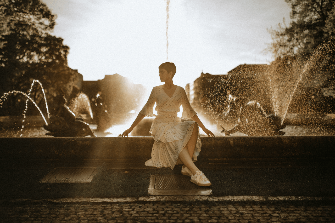A woman wearing gold jewelry posing by a water fountain.