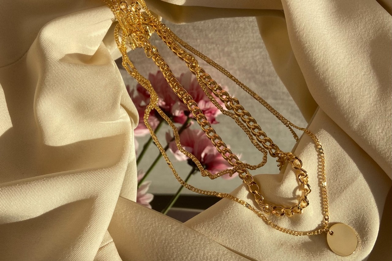 A group of necklaces sit messily on a mirror and tan fabric.