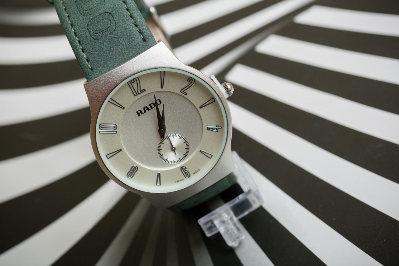 A retro Rado watch with a green leather strap sits on a striped surface.