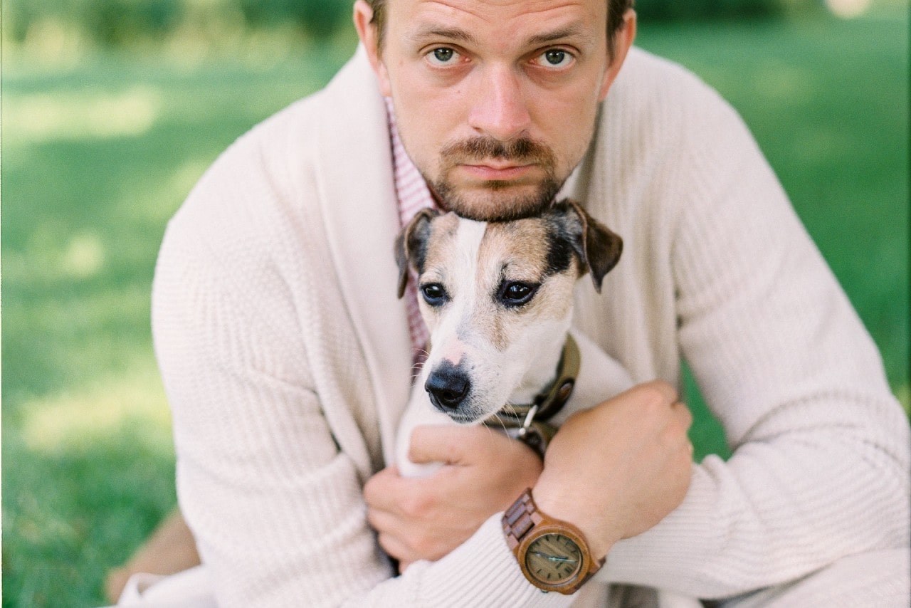A man hugs a terrier in a park, while wearing a wood grain watch.