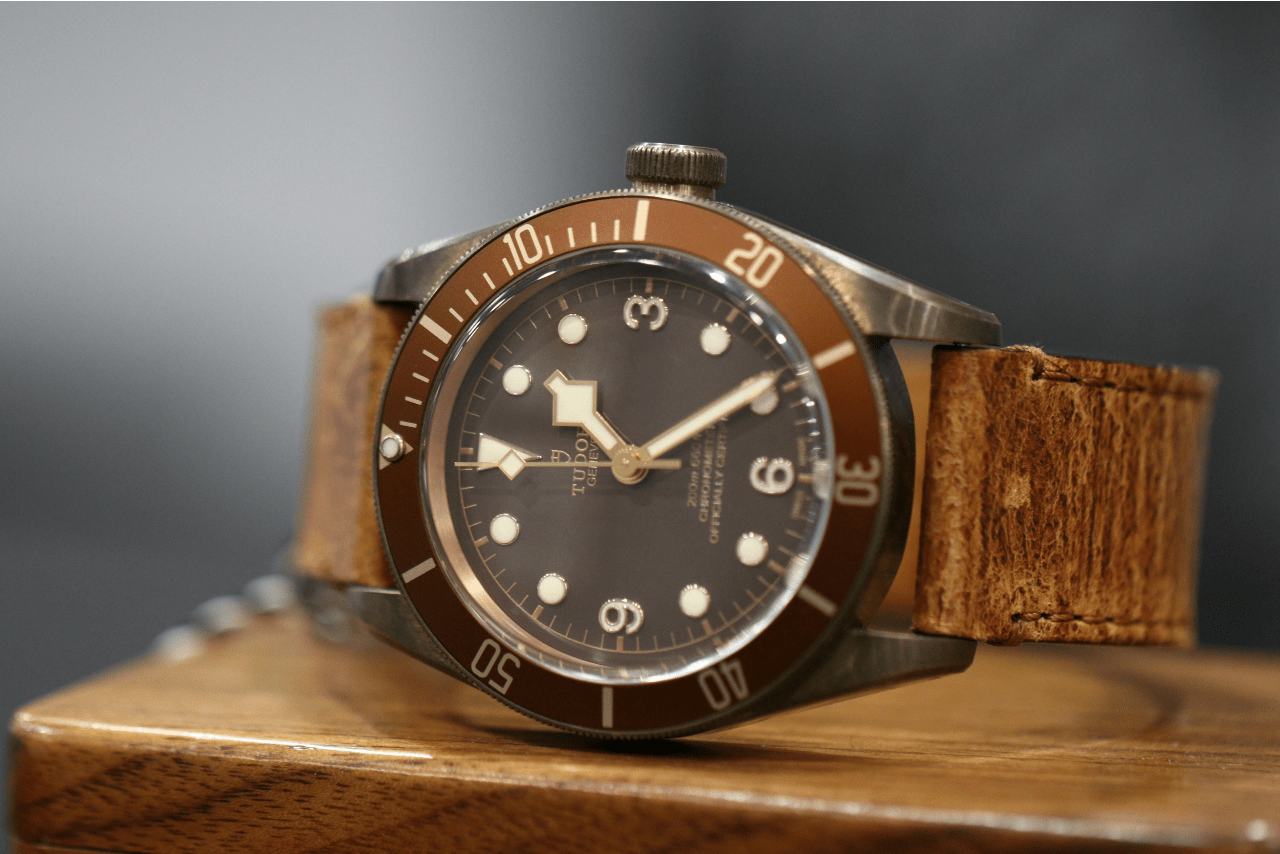 A close up of a TUDOR Geneve wristwatch with black dial and brown leather strap resting on a wooden surface. A close up of a TUDOR Geneve wristwatch with black dial and brown leather strap resting on a wooden surface.