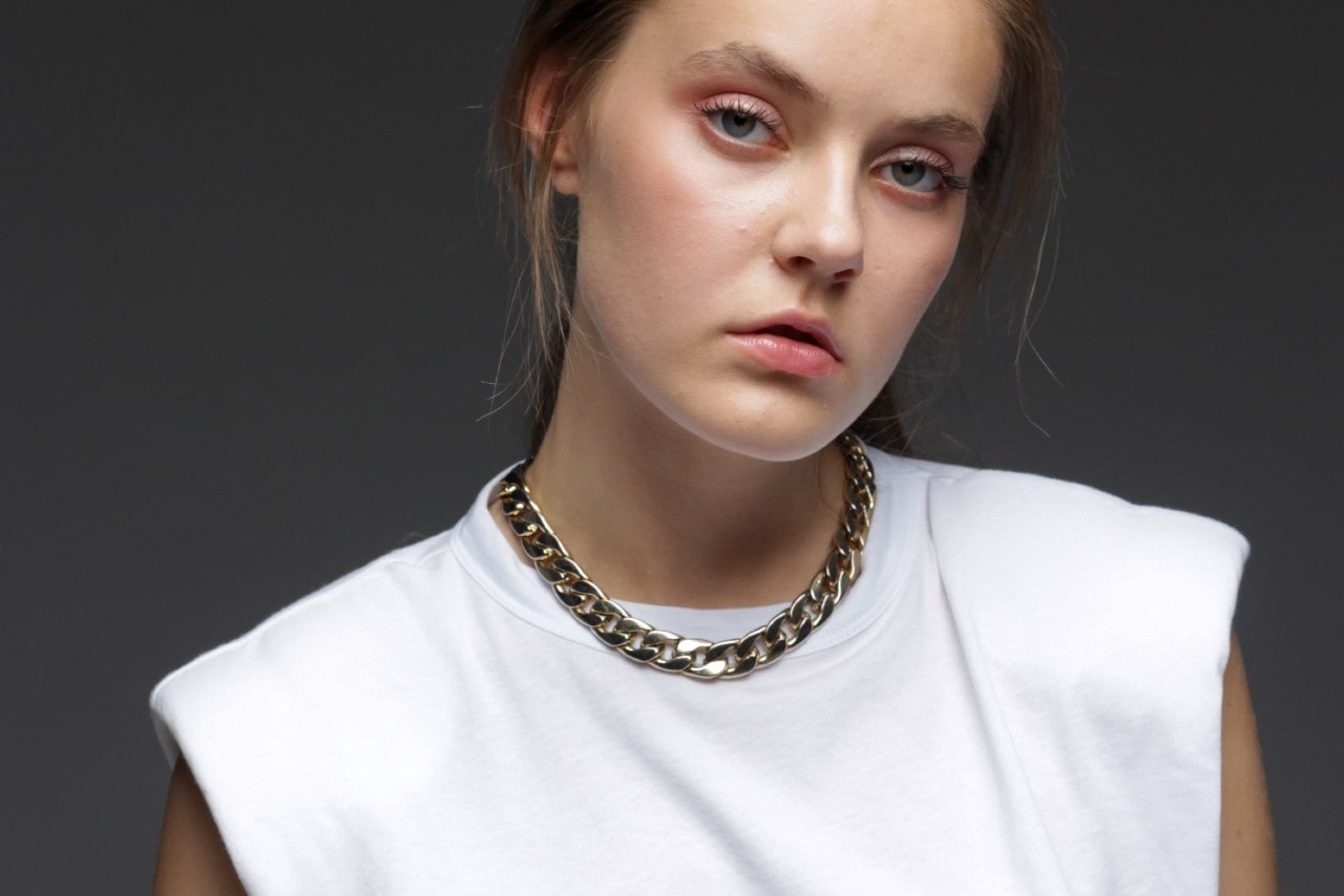 Young woman with a serene expression, wearing a white top and chunky gold chain necklace, against a dark background. Young woman with a serene expression, wearing a white top and chunky gold chain necklace, against a dark background.