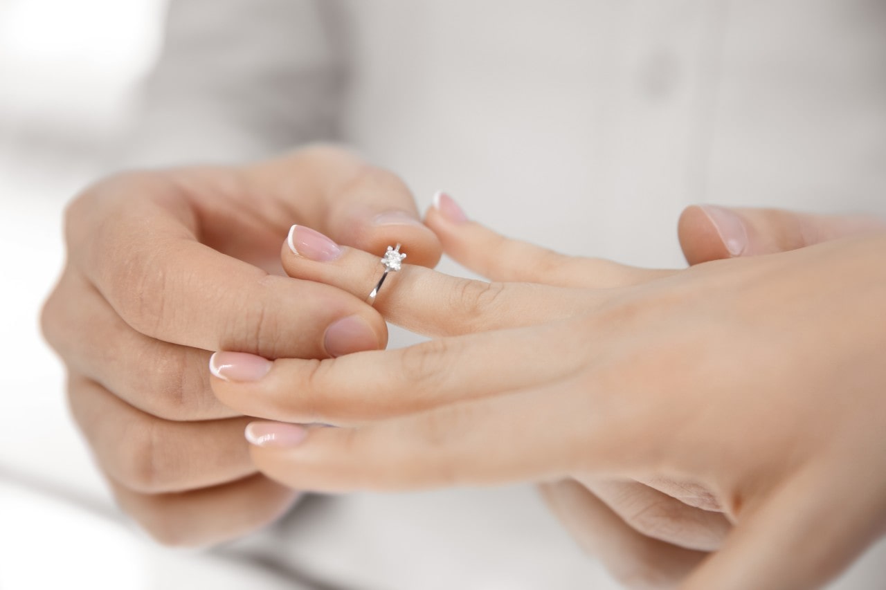 A person placing a solitaire diamond engagement ring on another’s finger. A person placing a solitaire diamond engagement ring on another’s finger.