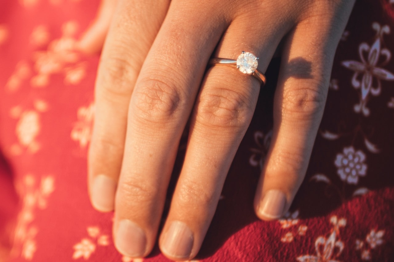 A close-up of a rose gold round cut diamond engagement ring on a person’s hand against a patterned red fabric. A close-up of a rose gold round cut diamond engagement ring on a person’s hand against a patterned red fabric.
