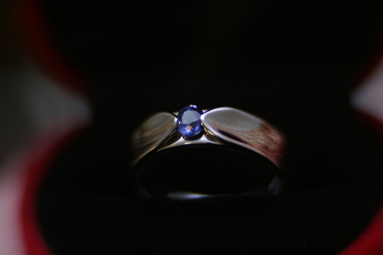 A close up of a silver-toned wedding band with an oval cut sapphire center stone against a dark backdrop.