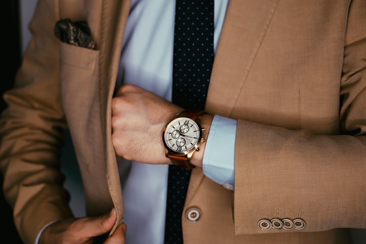 A man wearing a tan suit adjusts his jacket’s breast pocket while sporting a designer watch