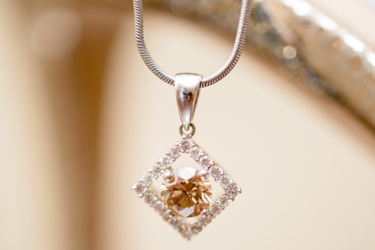 A close-up of a silver pendant necklace featuring a square diamond-studded frame with a sparkling champagne-colored gemstone at its center on a blurred background.