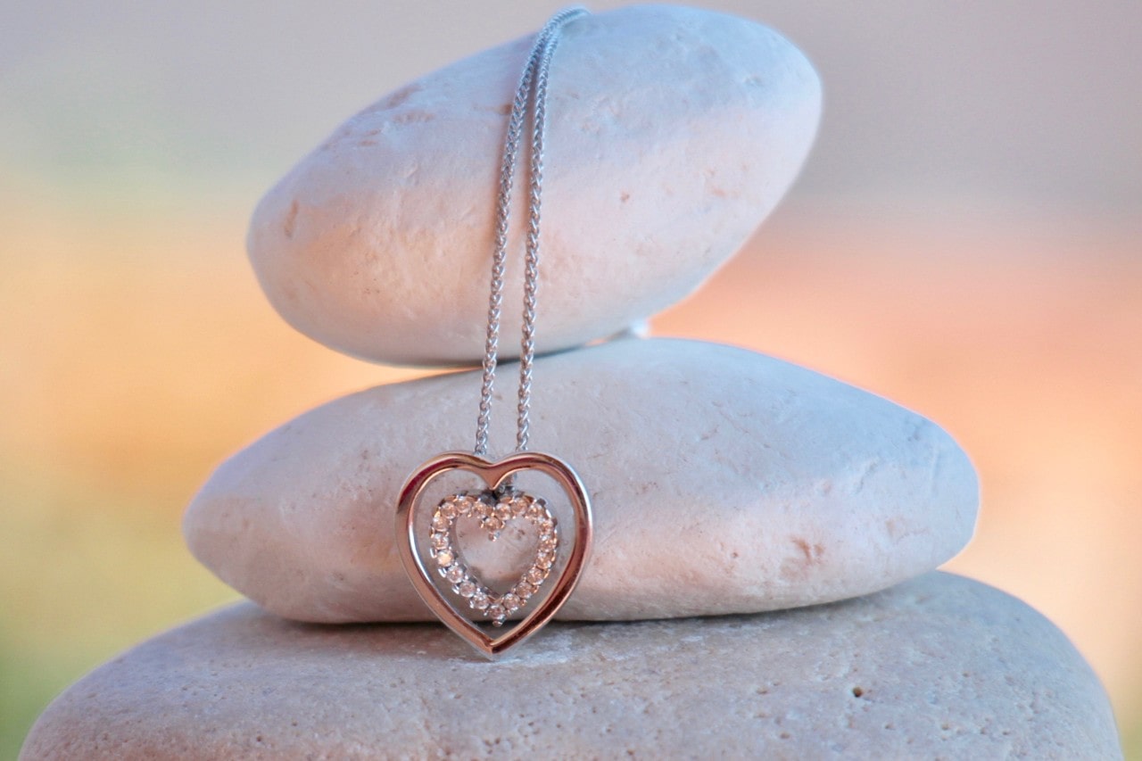 A gold rose silver heart-shaped pendant with an inner ring of sparkling diamonds hangs from a fine chain, displayed against smooth white stones