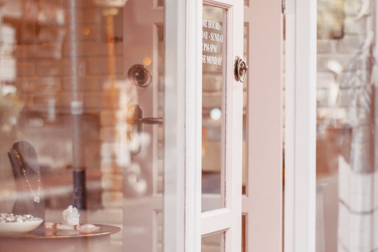 An exterior view of a small jewelry storefront with a small but elegant window display, the front door welcomingly ajar.