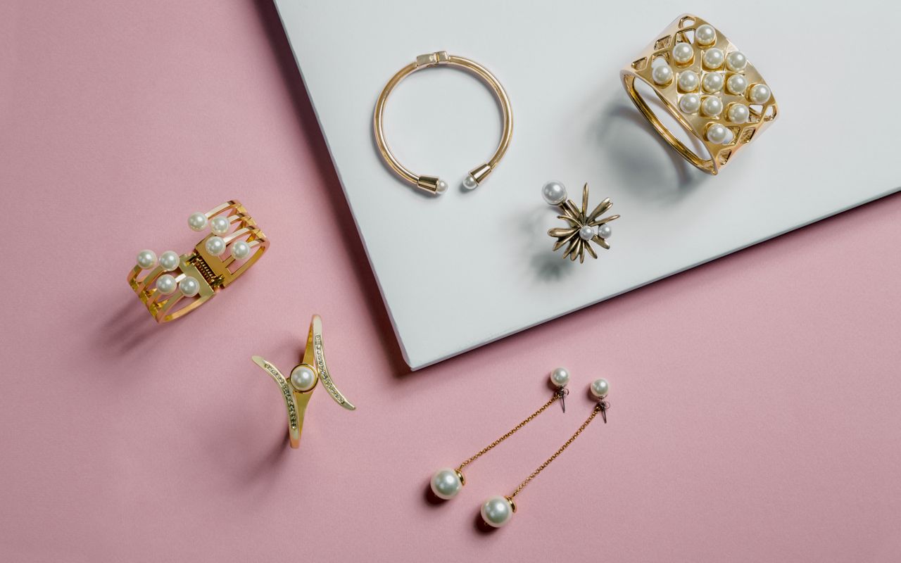 An elegant selection of gold jewelry with pearls on a pink backdrop and white surface.