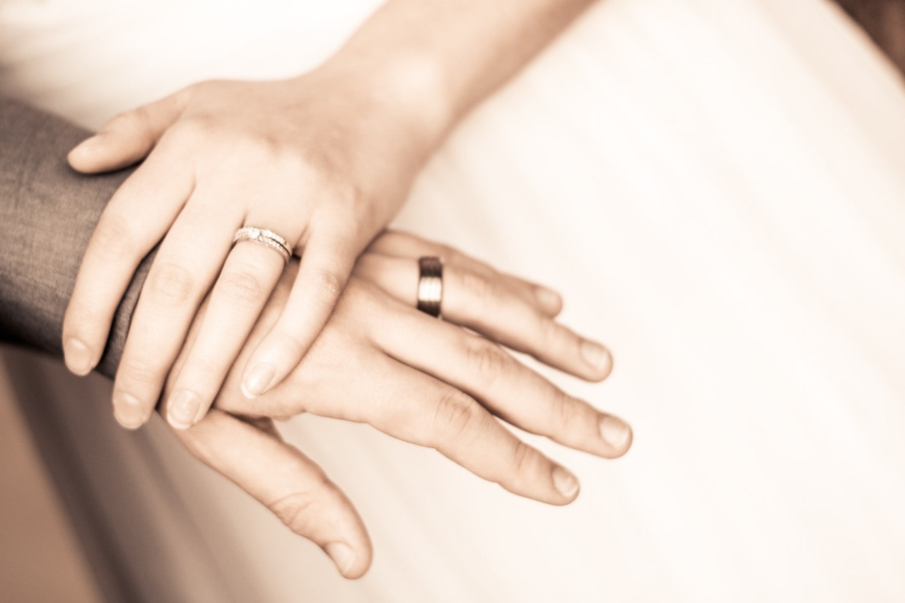 A couple’s hands together, showcasing his wedding band and brides’ stack against a white wedding gown.