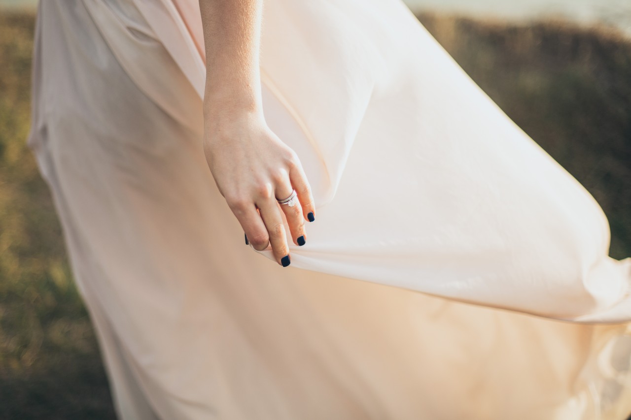 A bride holds her dress against the wind, showcasing a white gold diamond bridal stack against a sunlight hill.