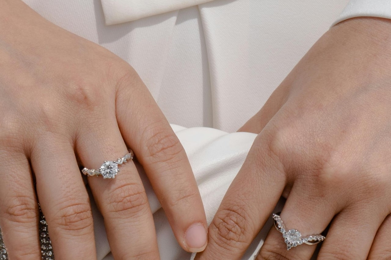 A close-up of a woman’s hands holding a white clutch, with two distinctive diamond rings on each of her middle fingers.