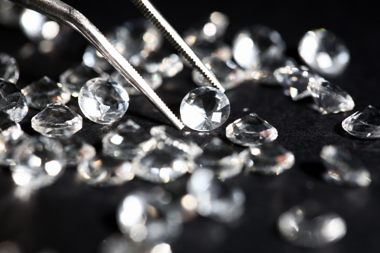 Close-up of a jeweler’s tweezers holding a round-cut diamond among many other loose diamonds on a dark surface.