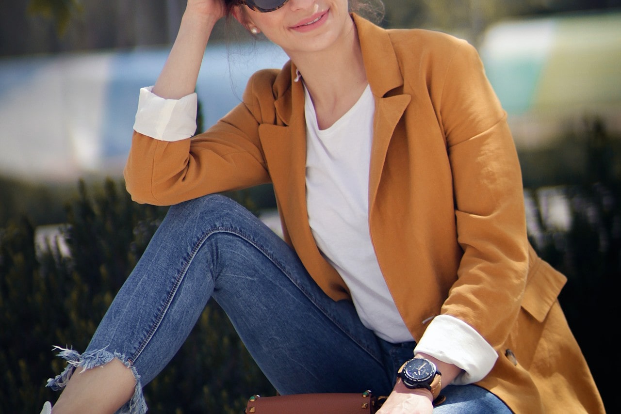 a woman in a burnt orange blazer and jeans smiling and wearing a black and brown watch