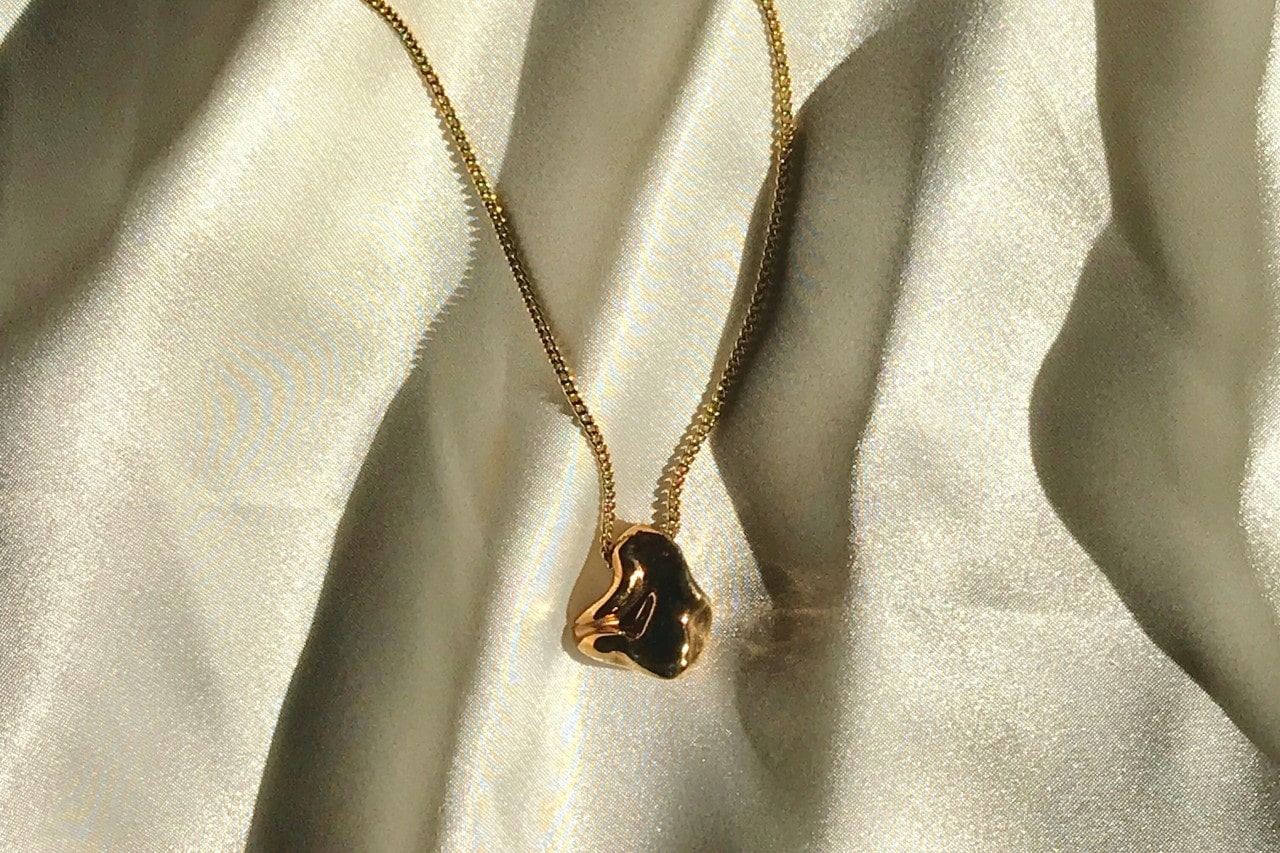 A yellow gold necklace with a sculptural pendant displayed atop a soft white sheet.