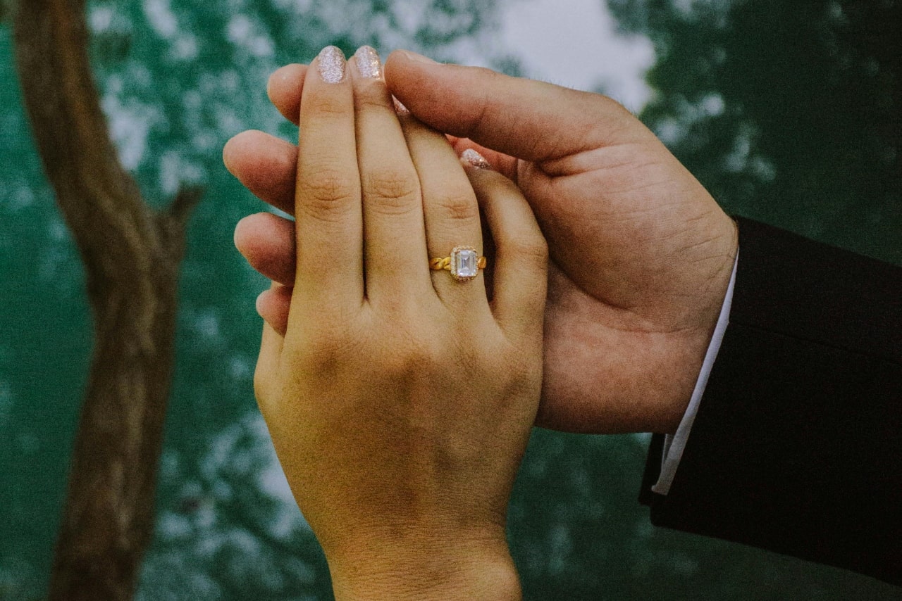 A close-up of a bride and groom holding hands, the bride's hand is facing the camera and showing off her mixed metal engagement ring.