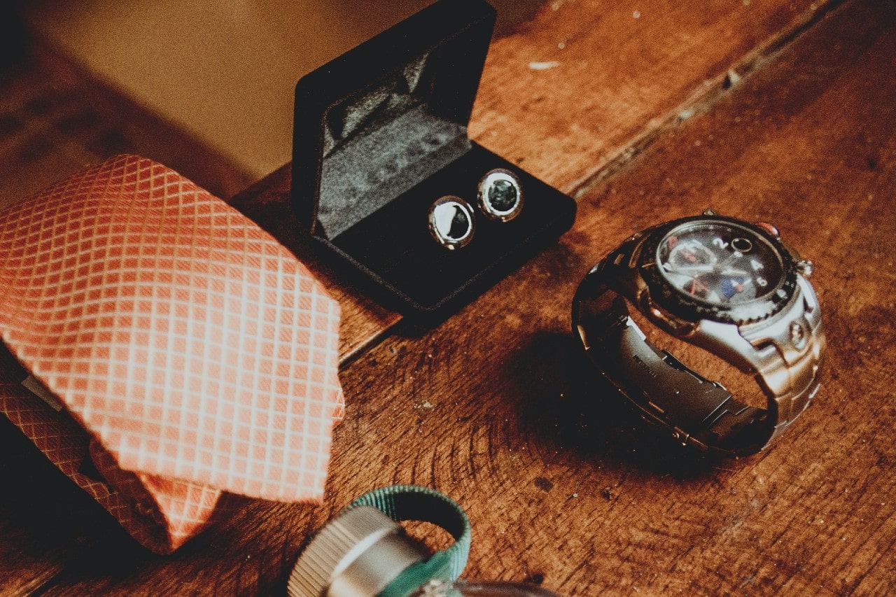 A silver luxury watch on a wooden table, next to cuff links and a tie