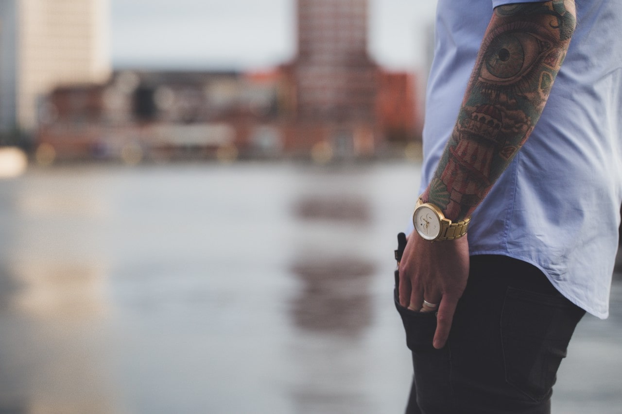 a man standing with his hand in his pocket, wearing a gold watch, with a cityscape in the background